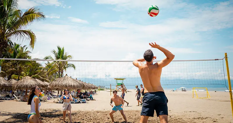 Man throws a volleyball on a beach with palm trees and people.