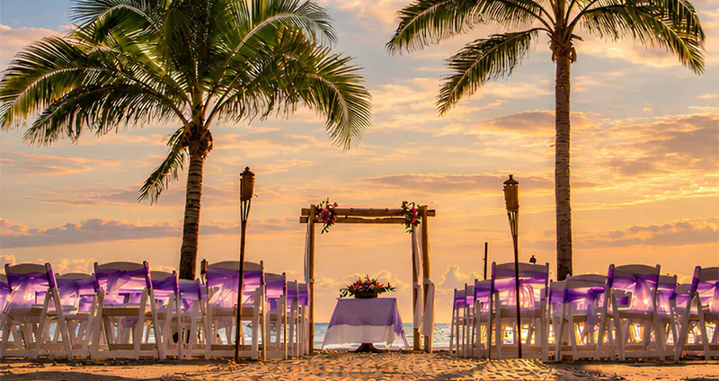 Beach weddings at sunset, white chairs and palm trees with decorated arch.