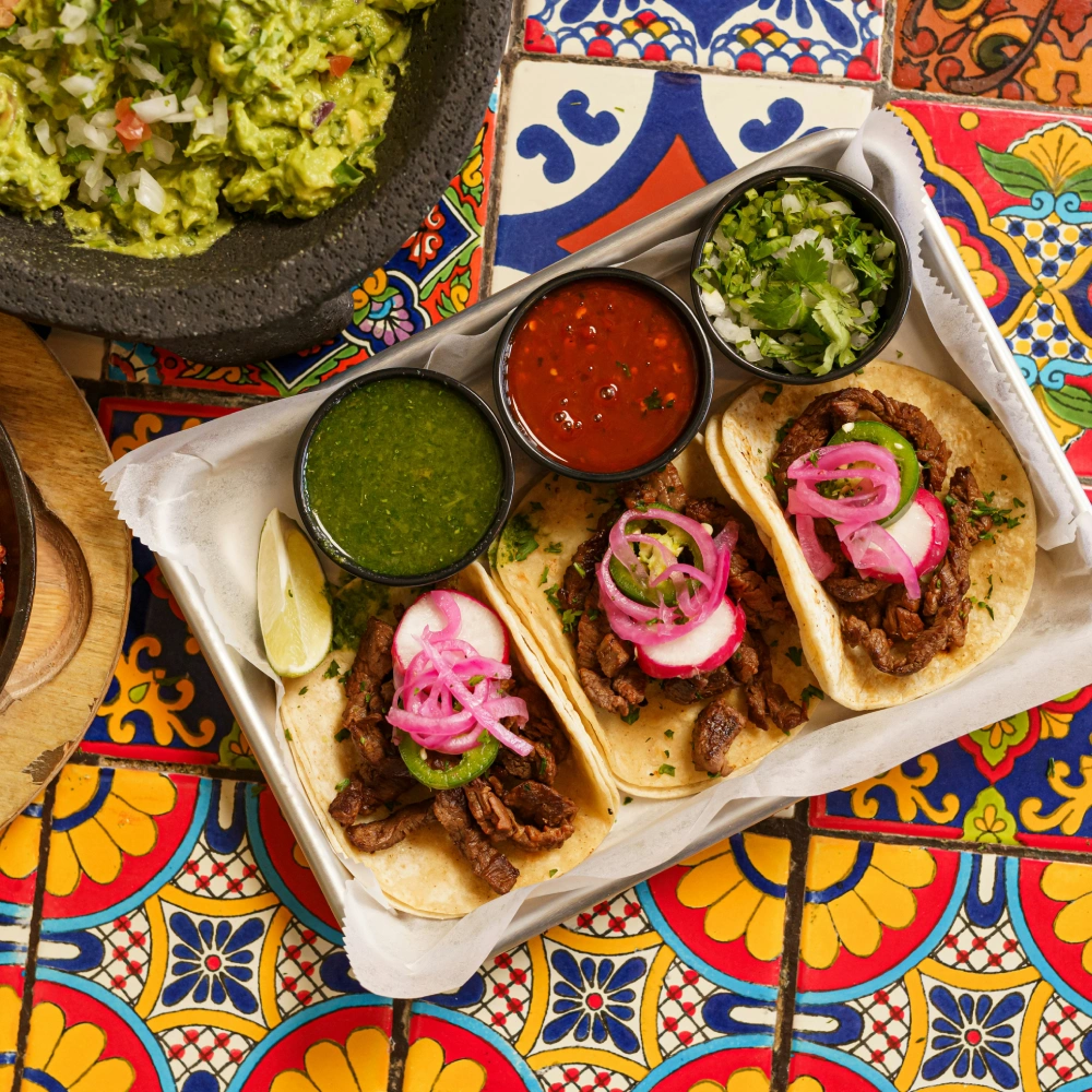 Tacos con carne, r&aacute;banos encurtidos, dos salsas y guacamole sobre fondo colorido