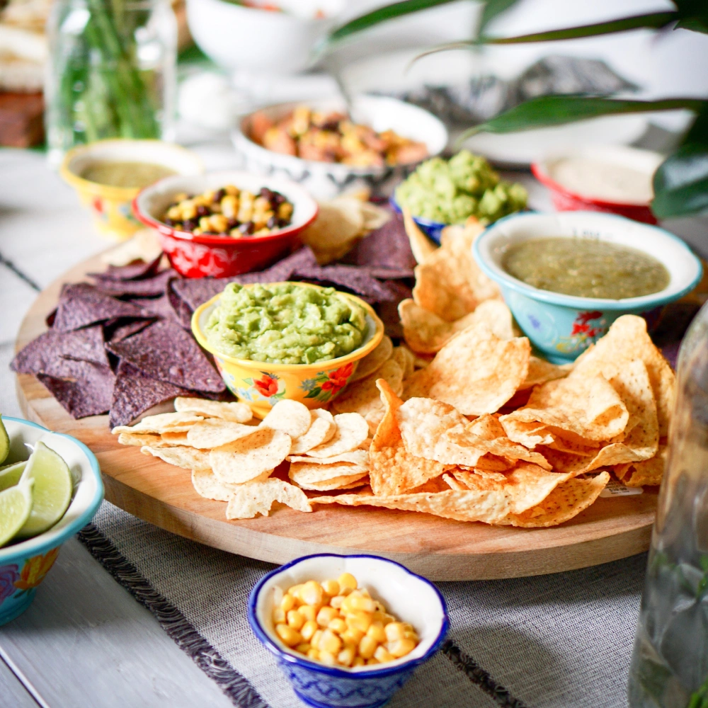 Table with tortilla chips, guacamole, beans, and sauces in bowls on wooden board