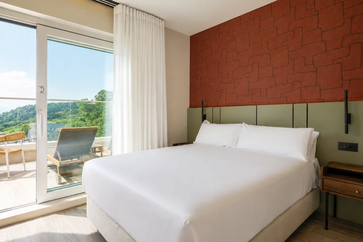 Habitaci&oacute;n moderna con cama blanca, cortinas y terraza con vistas verdes.