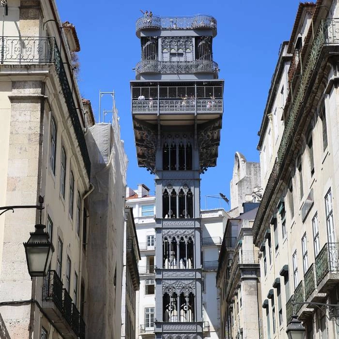 Torre estrecha ornamentada entre edificios antiguos, cielo azul.
