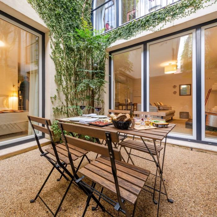 P&aacute;tio interno com mesa e cadeiras, janelas e plantas trepadeiras verdes.