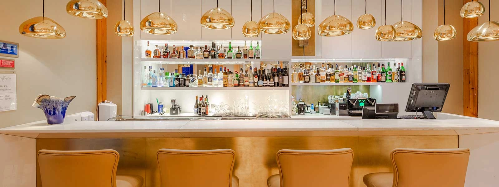 Bar moderno com balc&atilde;o, garrafas e lumin&aacute;rias suspensas douradas no interior.