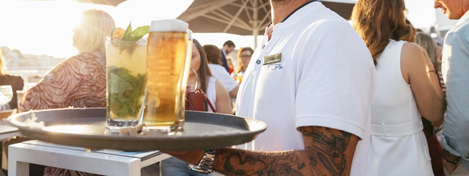 Gar&ccedil;om com tatuagem serve bebidas em evento ao ar livre, copos ao lado