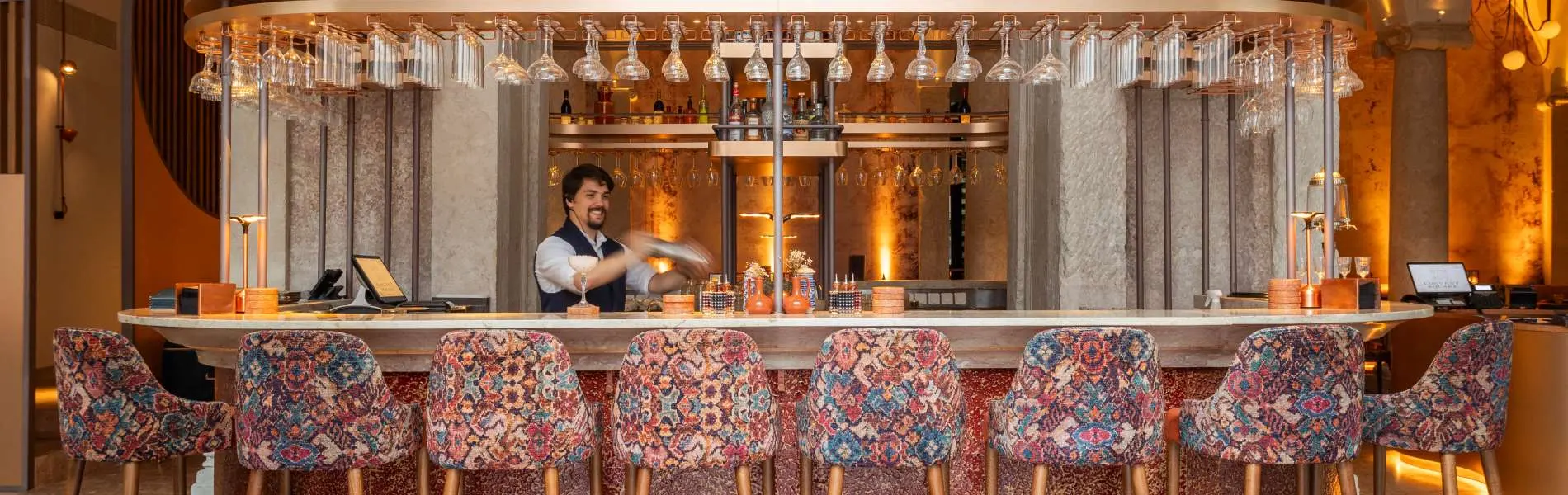 Barkeeper in einer beleuchteten Bar mit Tischen und einer Reihe von Gl&auml;sern und Flaschen im Hintergrund
