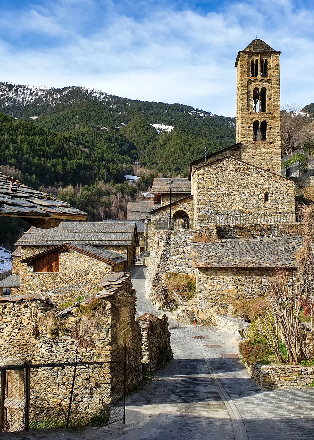 Iglesia de piedra con torre en un pueblo, rodeada de monta&ntilde;as y cielo azul