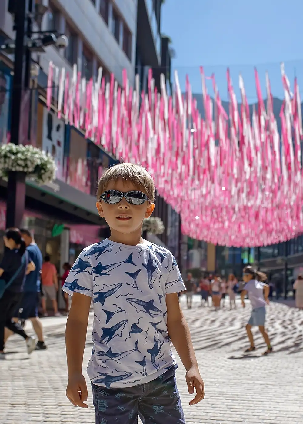 Ni&ntilde;o con gafas de sol, camiseta con tiburones, caminando en calle con banderines rosados