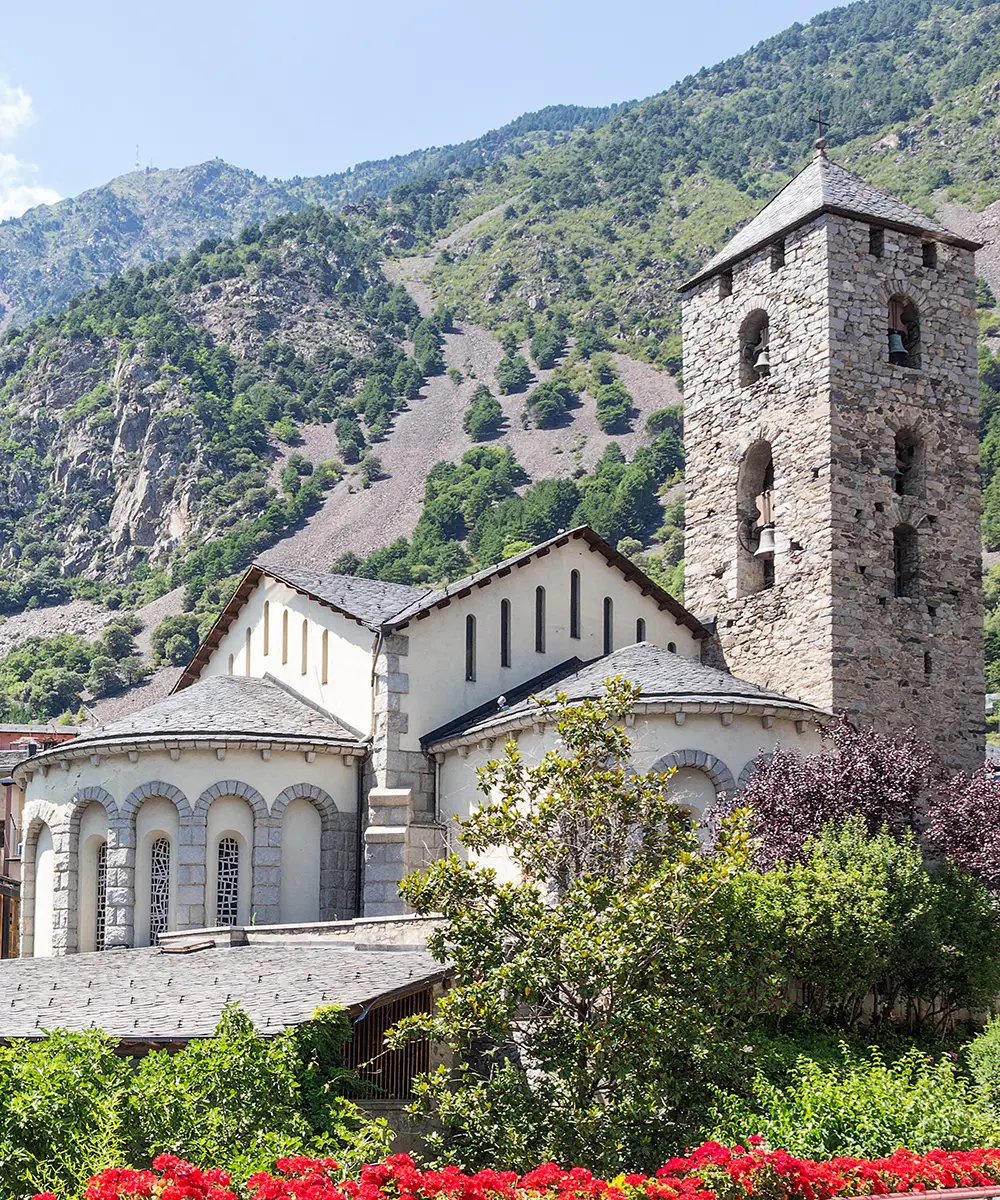 Iglesia de piedra y jard&iacute;n con flores rojas frente a monta&ntilde;as verdes y sendero