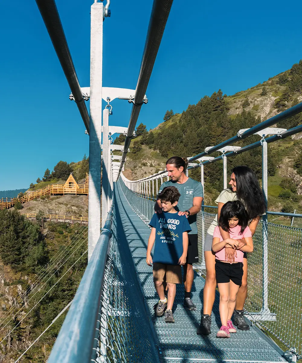 Familia caminando en puente colgante sobre un valle monta&ntilde;oso, cielo azul.
