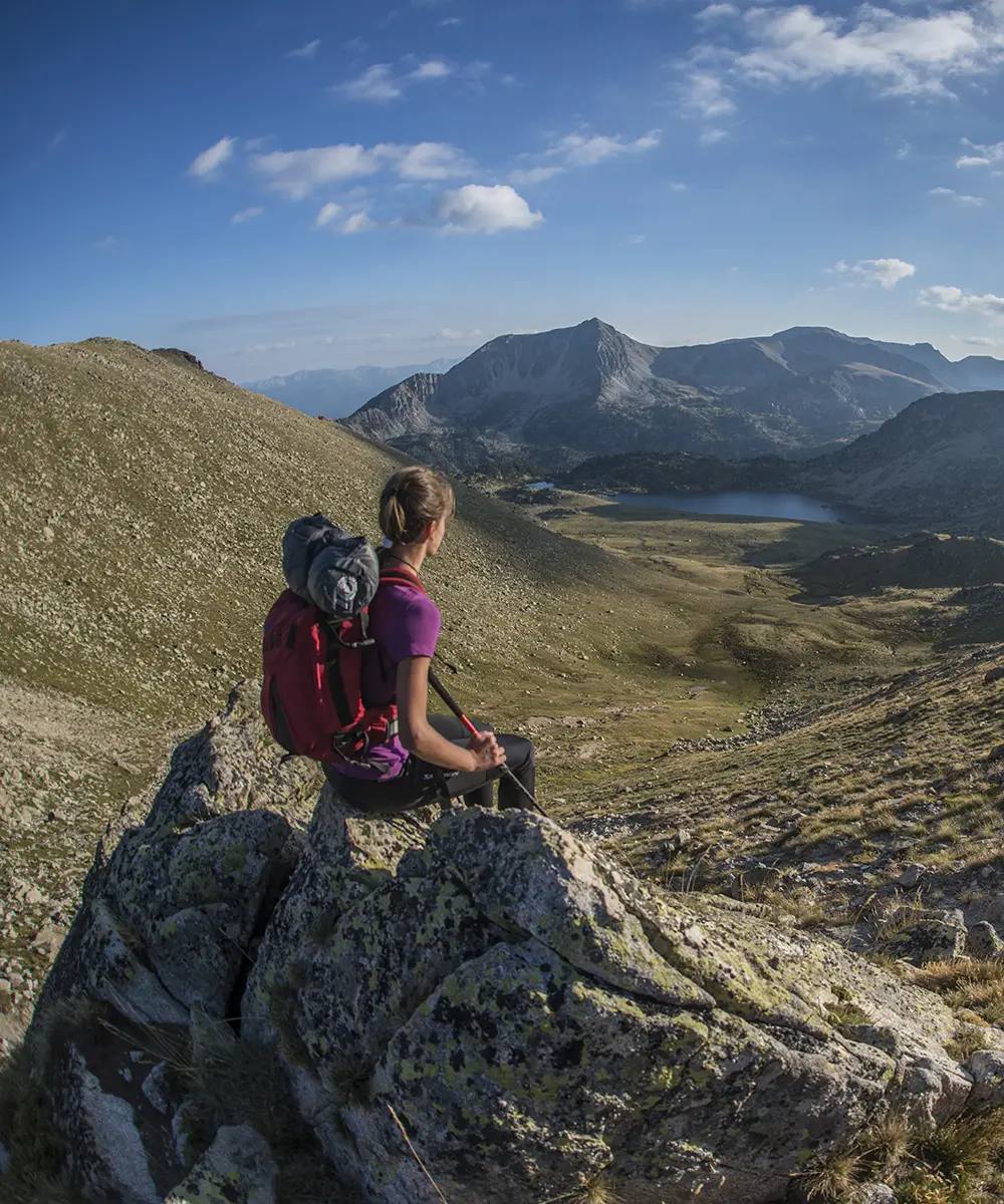 Senderista con mochila, sentada en la monta&ntilde;a, mirando un valle y un lago.