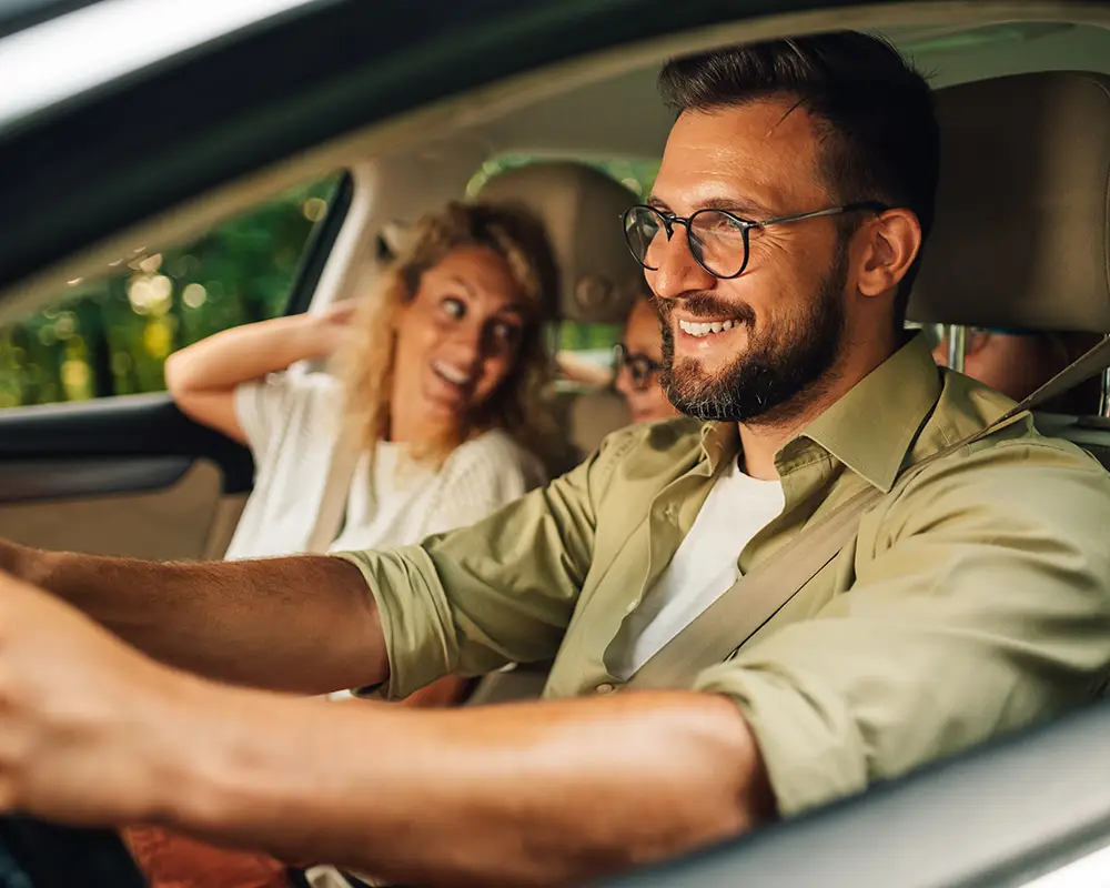 Hombre sonriente conduciendo; mujer y otra persona detr&aacute;s en el coche