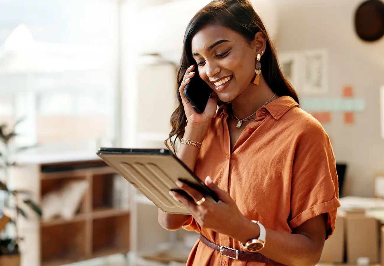 Mujer sonriente habla por tel&eacute;fono y revisa una tableta en oficina.