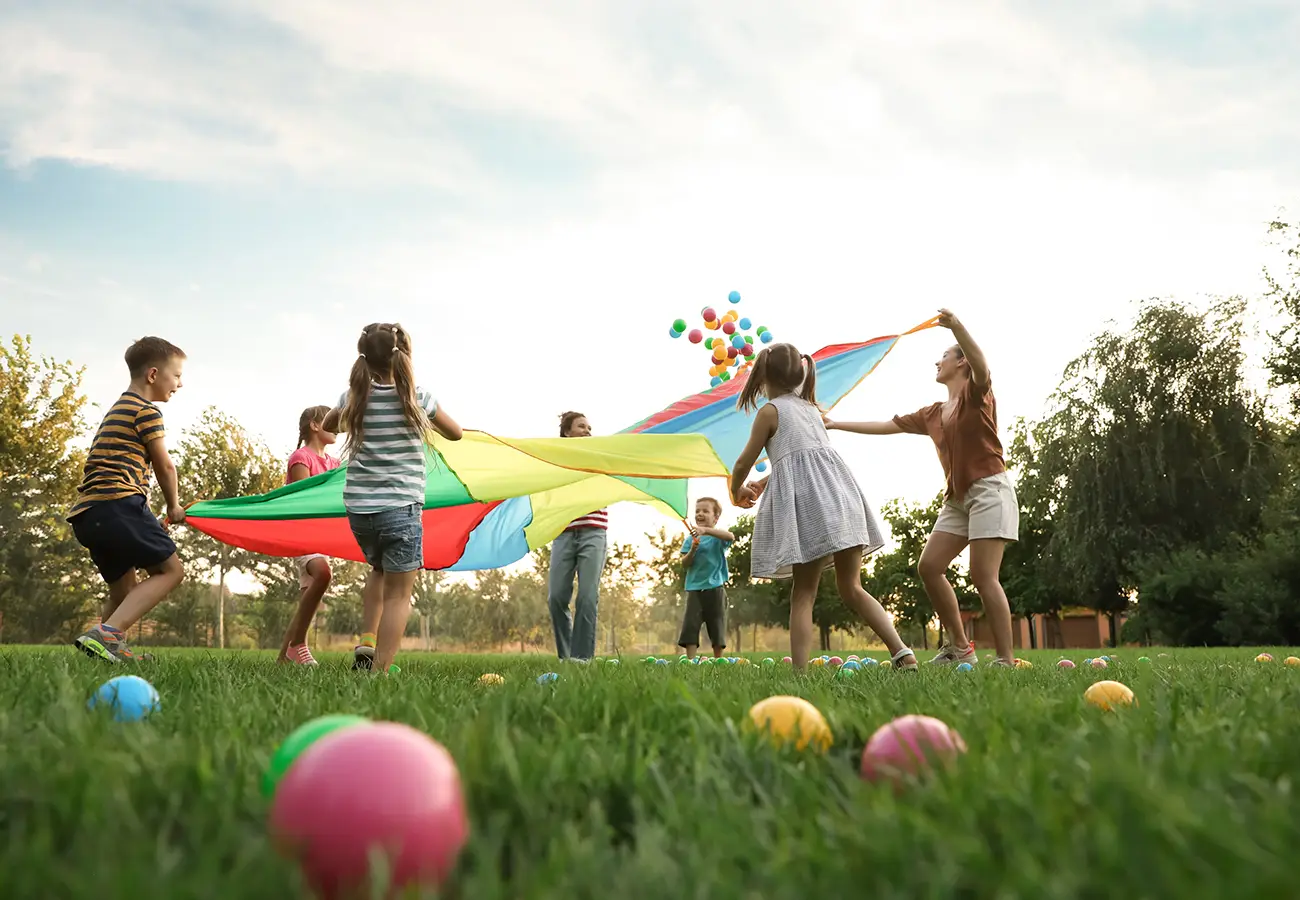 Ni&ntilde;os jugando con paraca&iacute;das multicolor y globos en el parque al atardecer