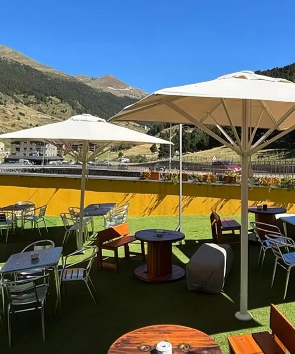 Terraza con mesas y sombrillas junto a un campo, rodeada de monta&ntilde;as.