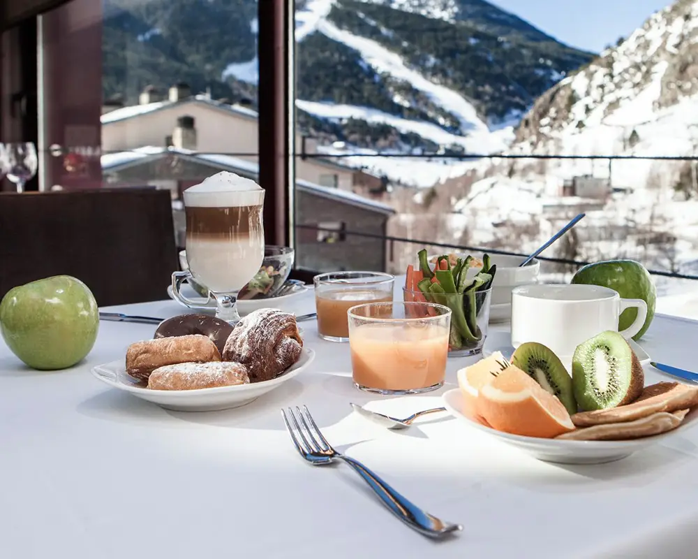 Desayuno en mesa con frutas y bebidas frente a monta&ntilde;as nevadas.