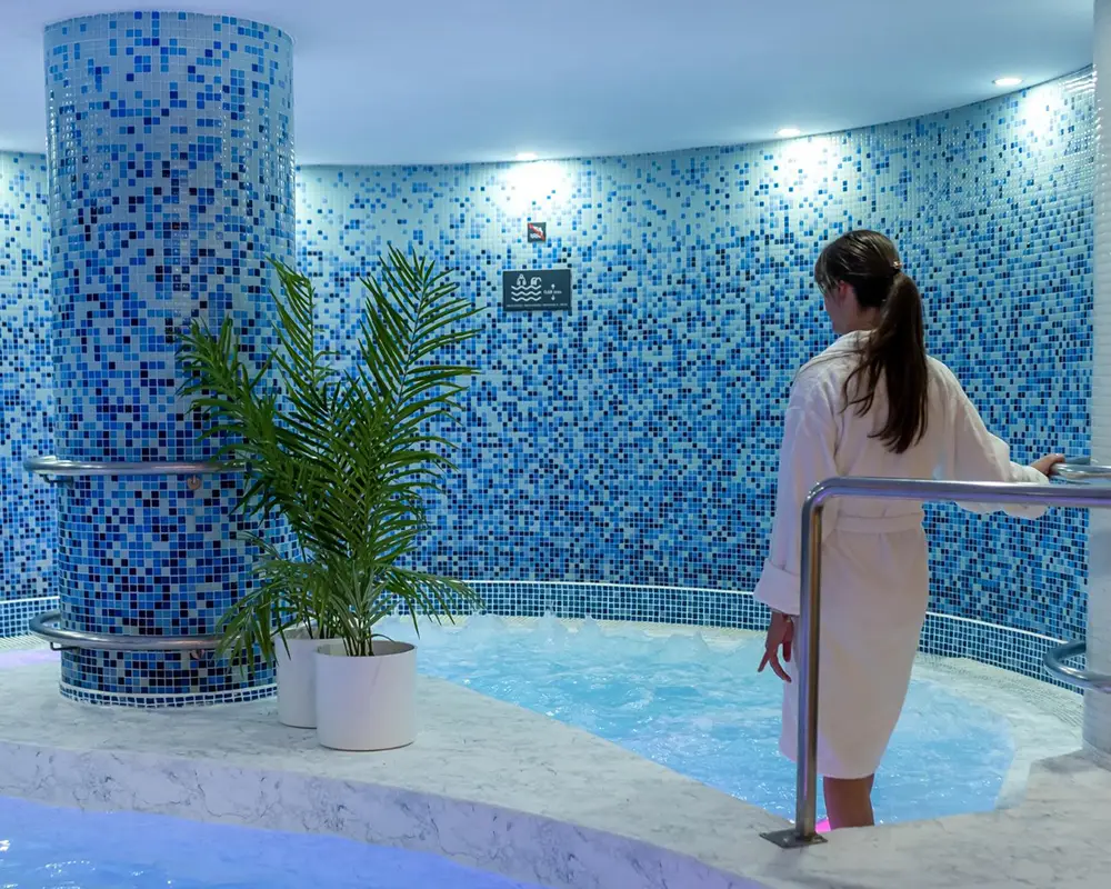 Mujer con bata en spa de mosaicos azules y jacuzzi con plantas