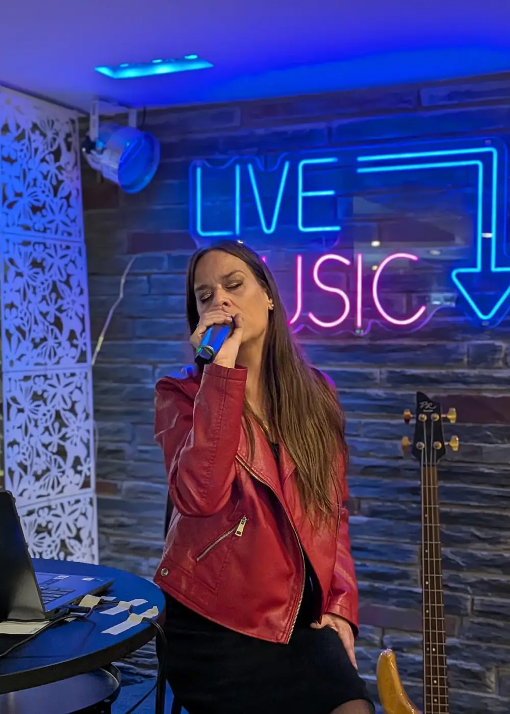 Cantante con micr&oacute;fono en escenario, chaqueta roja y luces ne&oacute;n &ldquo;LIVE MUSIC&rdquo;