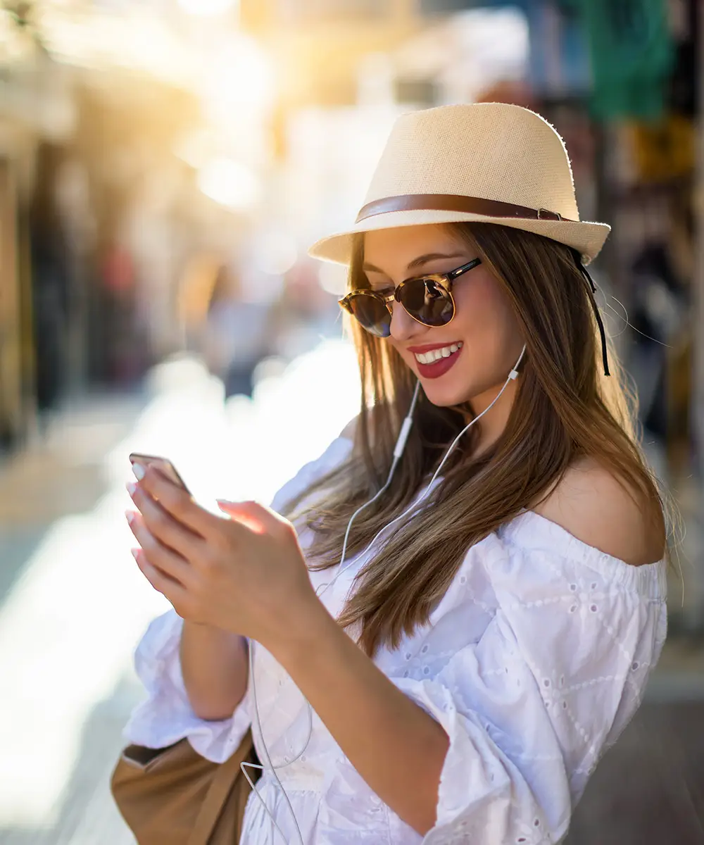 Mujer sonriente con sombrero, gafas y aud&iacute;fonos, usando el m&oacute;vil al aire libre.