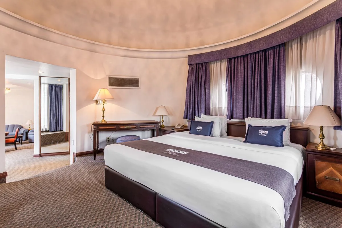 Dormitorio de hotel con cama doble, cortinas moradas y l&aacute;mparas de mesa