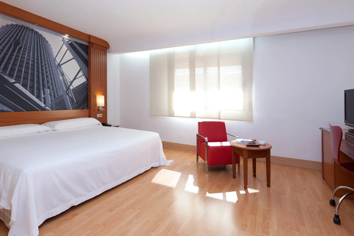 Bright hotel room with bed, red armchair, and desk next to the window.