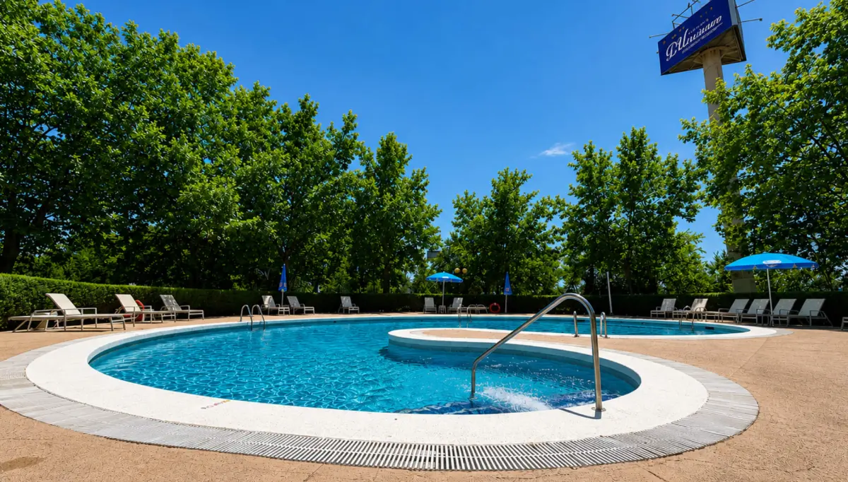 Piscina exterior con tumbonas, sombrillas y cielo azul, rodeada de &aacute;rboles.