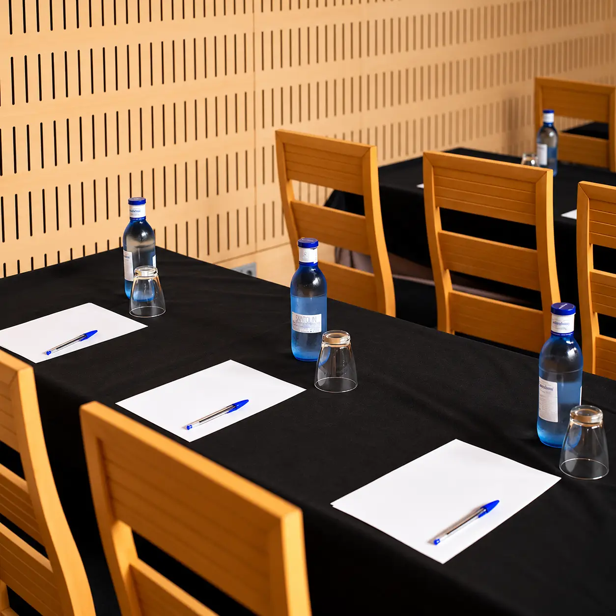Mesa con manteles negros, botellas de agua y sobres blancos con bol&iacute;grafos azules.