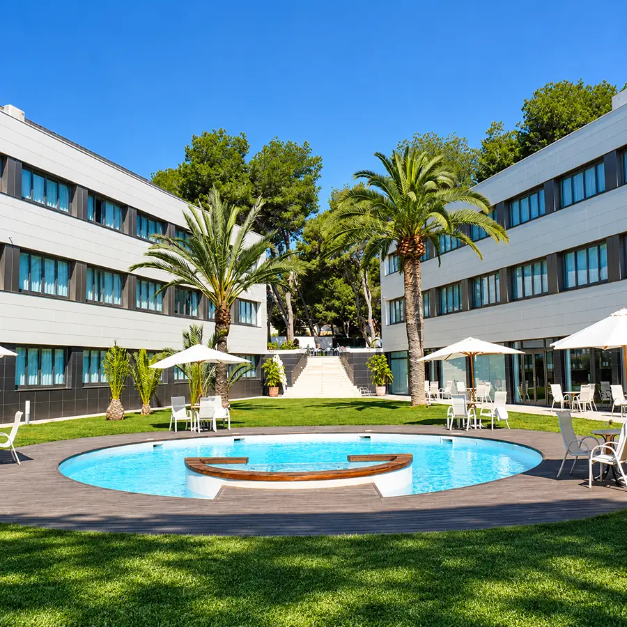 Piscina circular en jard&iacute;n con palmeras y edificios de hotel bajo cielo azul