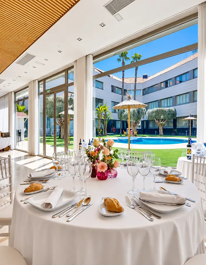 Mesa servida con copas y flores, vista a piscina y jard&iacute;n desde un sal&oacute;n.
