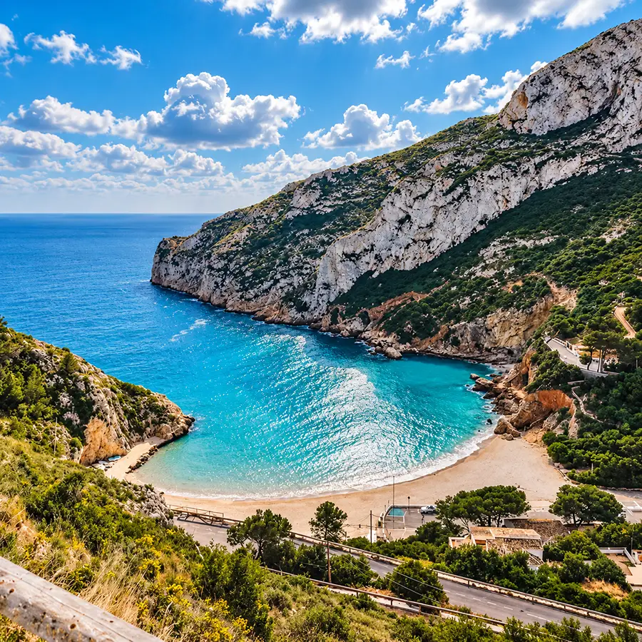 Playa de aguas turquesa entre acantilados, con camino y vegetaci&oacute;n costera.
