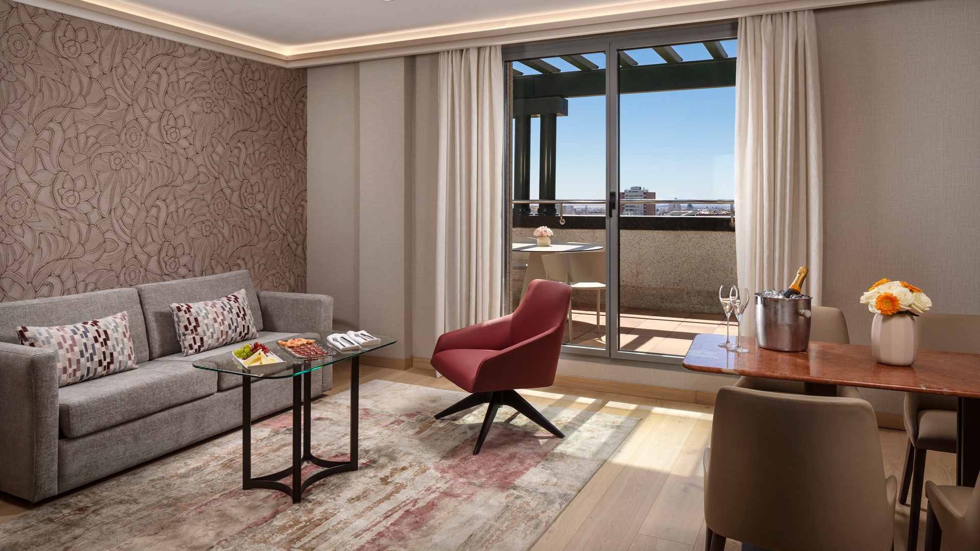 Sala moderna con sof&aacute;, silla roja y puerta a un balc&oacute;n soleado.