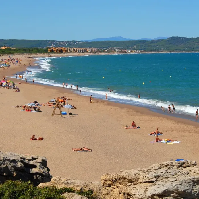 Playa soleada con personas tomando el sol y nadando en el mar azul, montañas al fondo.