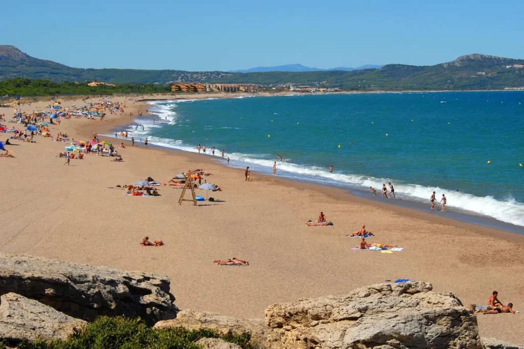 Playa concurrida con bañistas, sombrillas y mar azul. Montañas al fondo, cielo despejado.