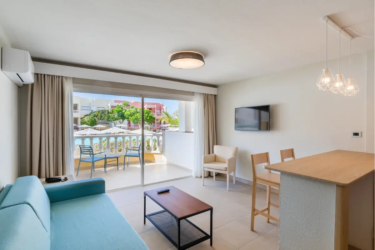 Sala moderna con sof&aacute; azul, balc&oacute;n, televisor y mesas en madera clara.