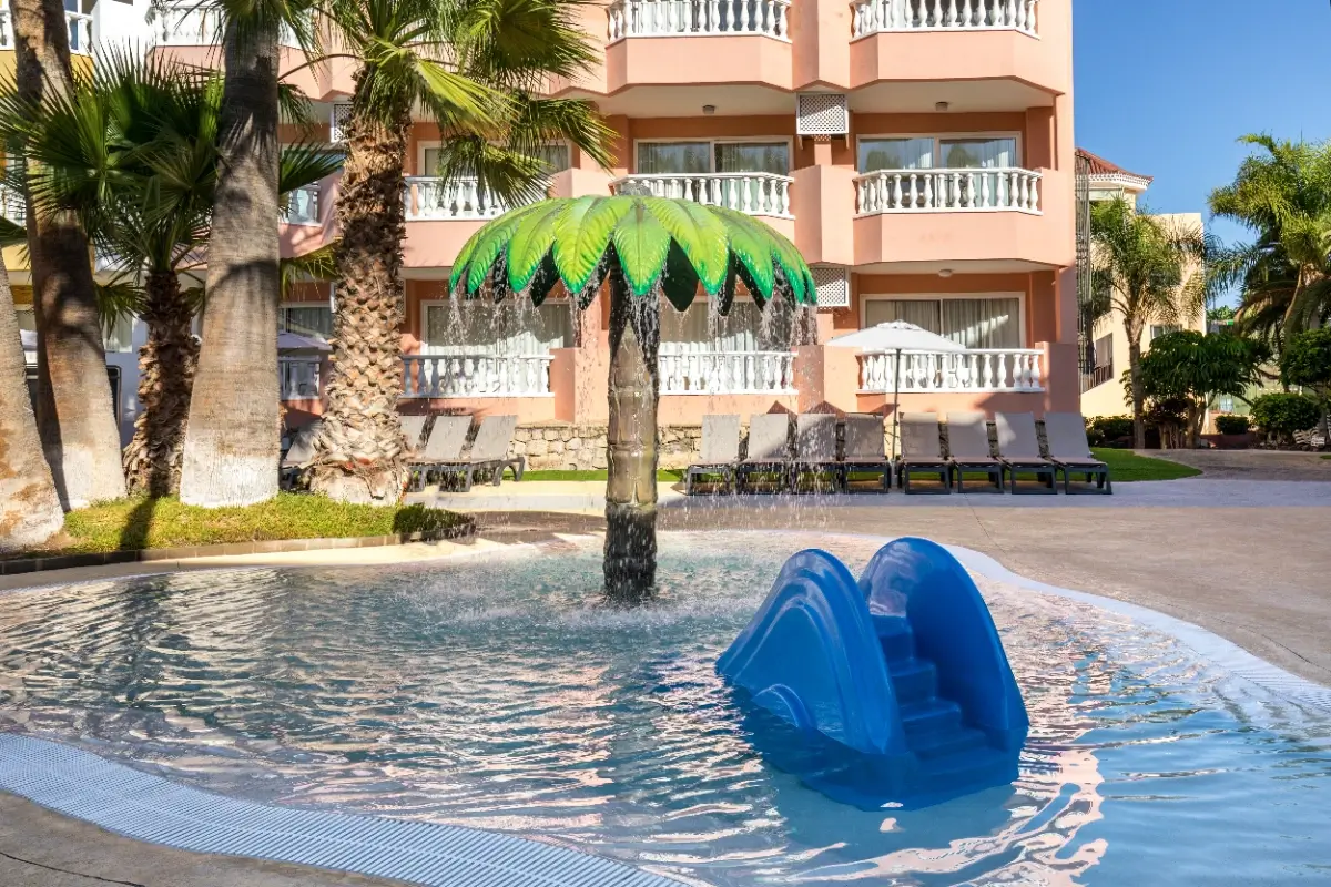 Piscina infantil con tobog&aacute;n azul y palmera en un hotel tropical.