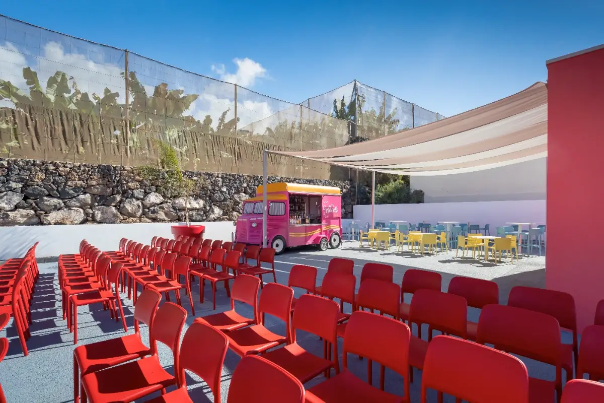 Sillas rojas al aire libre, food truck y mesas multicolores bajo toldo.