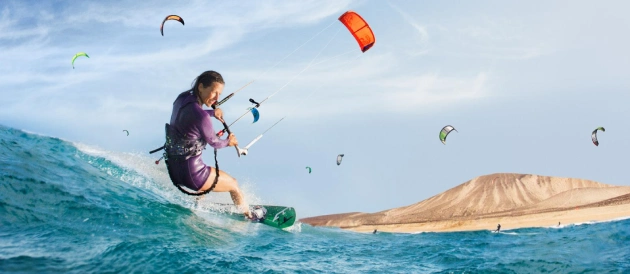 Persona practicando kitesurf en el mar, con cerros y varias cometas al fondo.