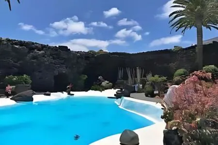 Piscina rodeada de palmeras y rocas bajo un cielo azul con nubes.