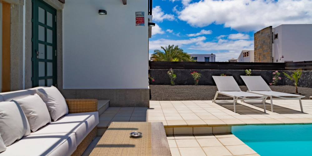 Patio con piscina, tumbonas y sofá blanco bajo un cielo parcialmente nublado.