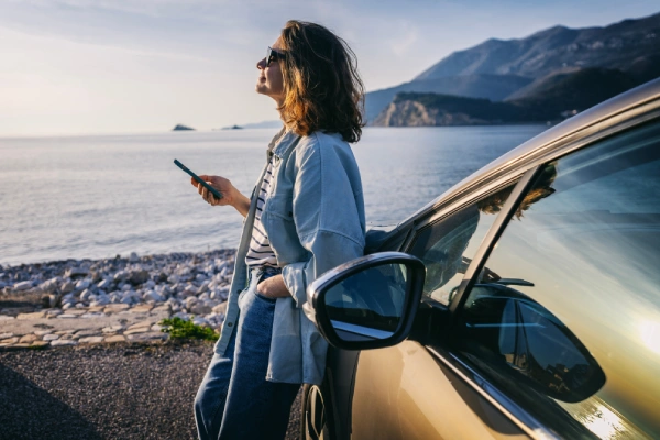 Mujer con móvil se relaja junto a un coche frente al mar y las montañas.