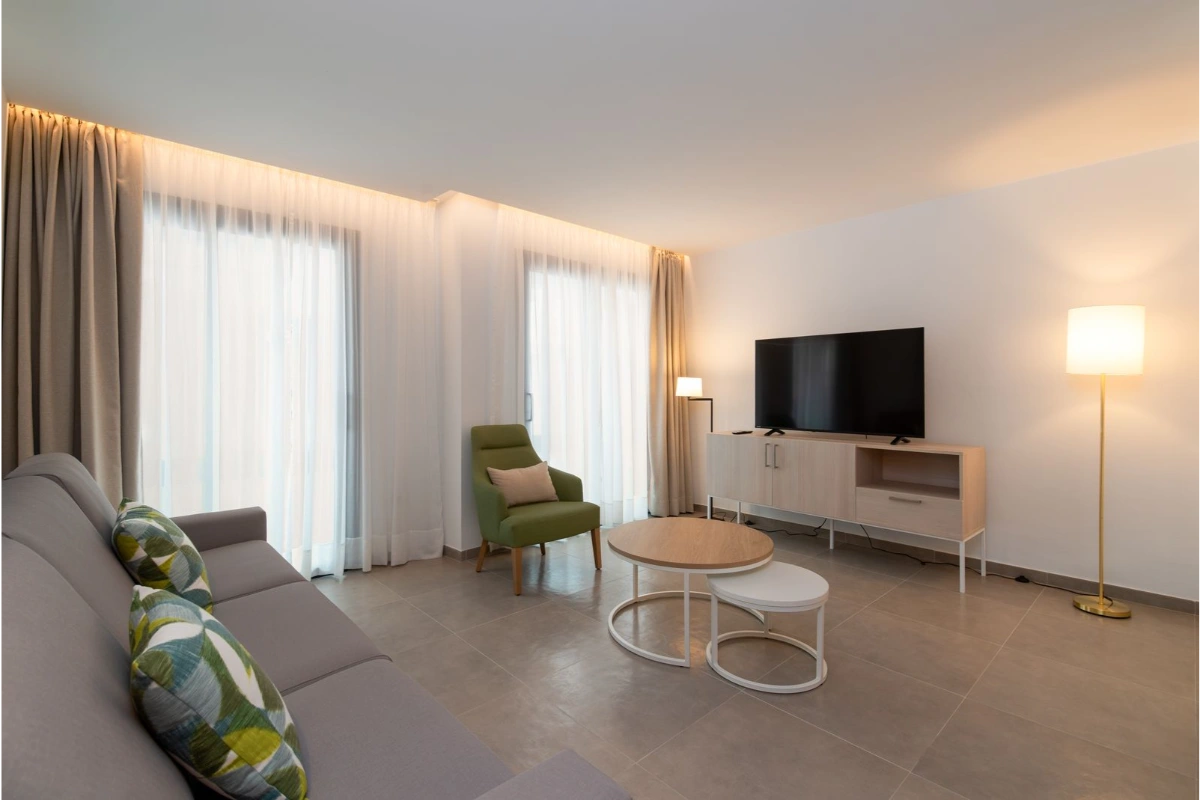 Sala moderna con sof&aacute; gris, sill&oacute;n verde, TV y mesas redondas.