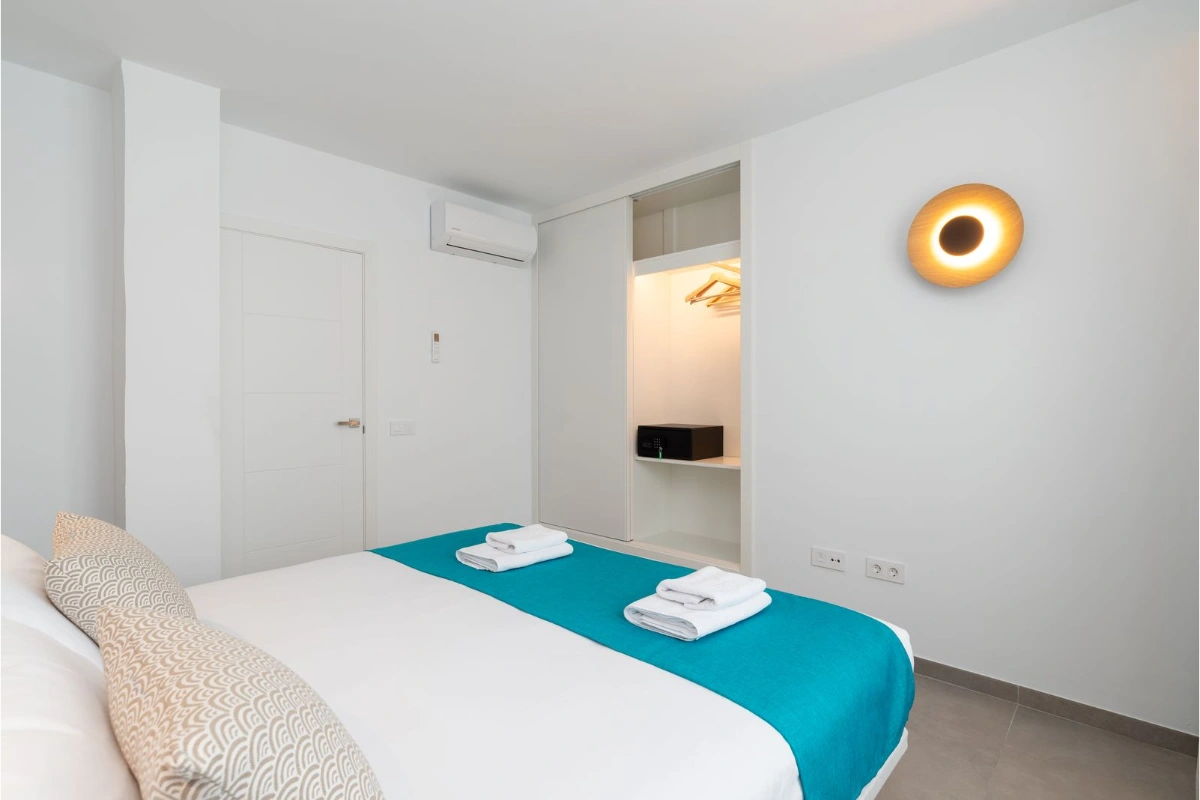 Habitaci&oacute;n moderna con cama blanca, cobertor turquesa y luz c&aacute;lida en la pared.
