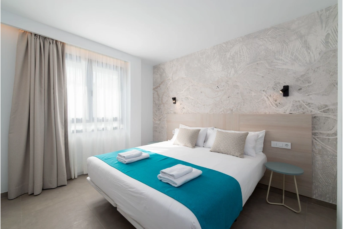 Cama doble con cojines y toallas, junto a ventana con cortinas en habitaci&oacute;n moderna.