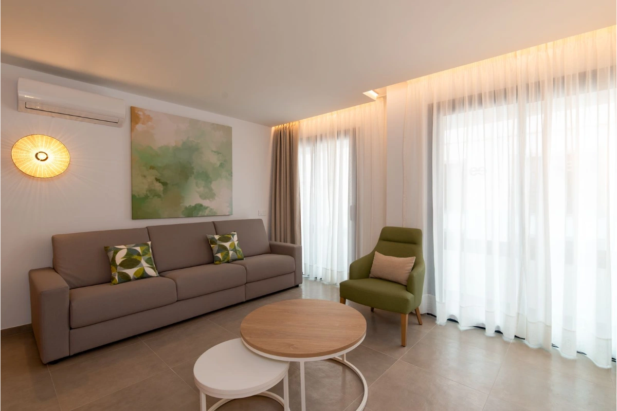 Sala moderna con sof&aacute;, sill&oacute;n verde y mesa redonda, luz natural entrando por ventanas.