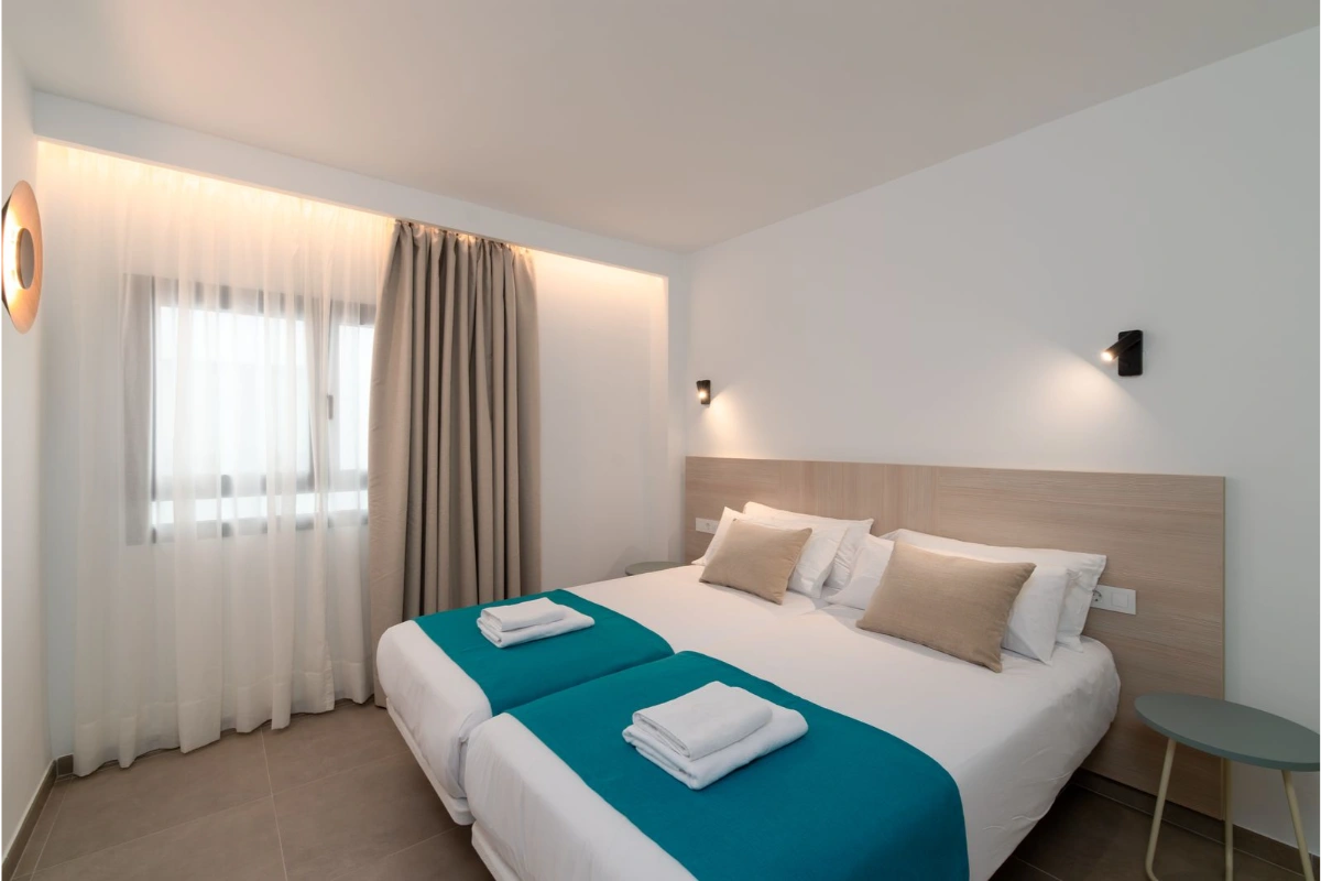 Habitaci&oacute;n moderna con cama doble, edred&oacute;n turquesa y mesa auxiliar.