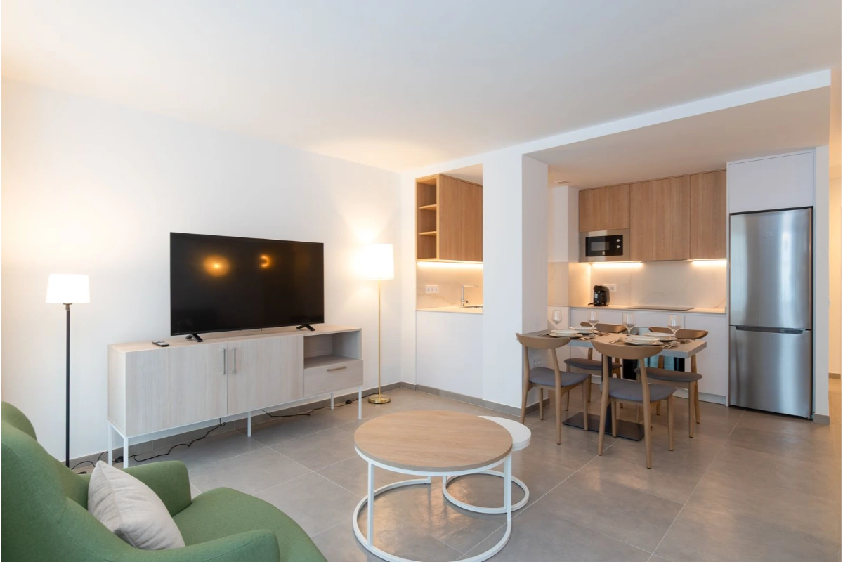 Sala y cocina moderna con muebles claros y televisi&oacute;n grande en la pared.