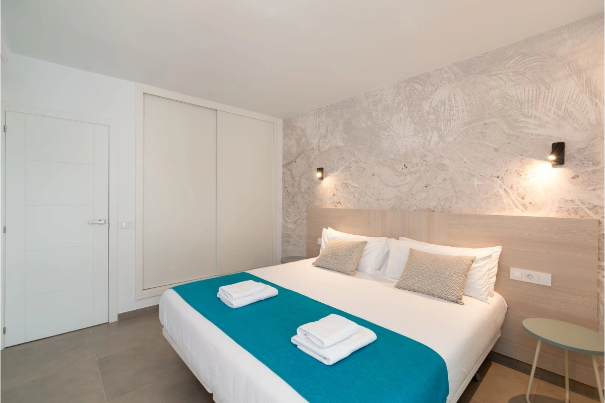 Cama doble con s&aacute;banas blancas y manta azul, en habitaci&oacute;n moderna.