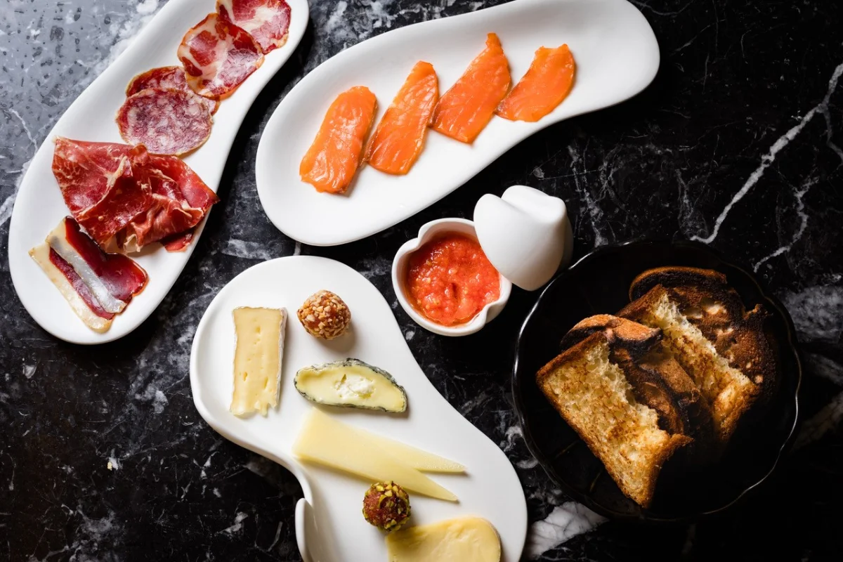 Platos con embutidos, salmón, quesos y tostadas sobre mesa de mármol.