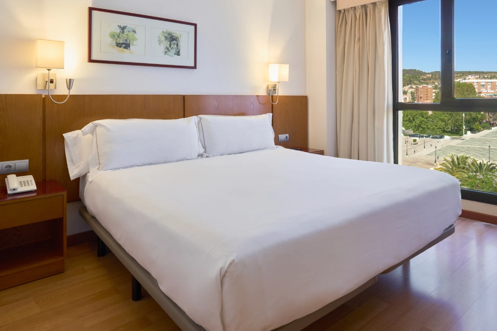 Dormitorio moderno con cama blanca y ventana con vista exterior.