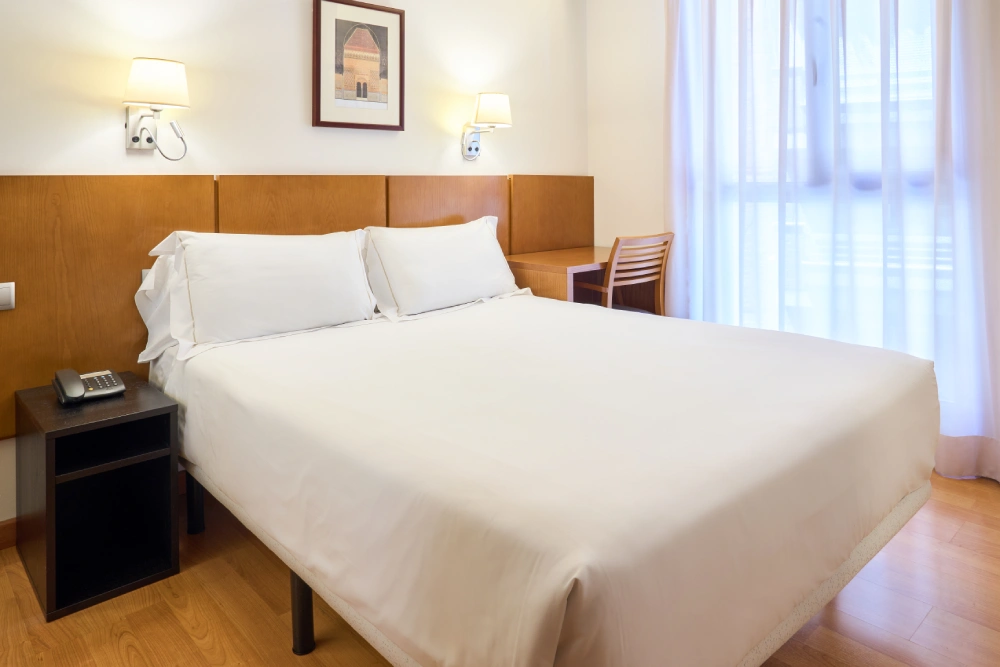 Habitaci&oacute;n de hotel con cama doble, mesita y cortinas junto a la ventana.